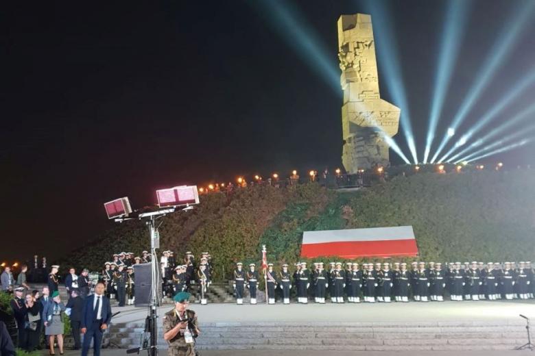 Oświetlony pomnik, pod pomnikiem duża flaga Polski oraz ustawieni w rzędach żołnierze marynarki wojennej