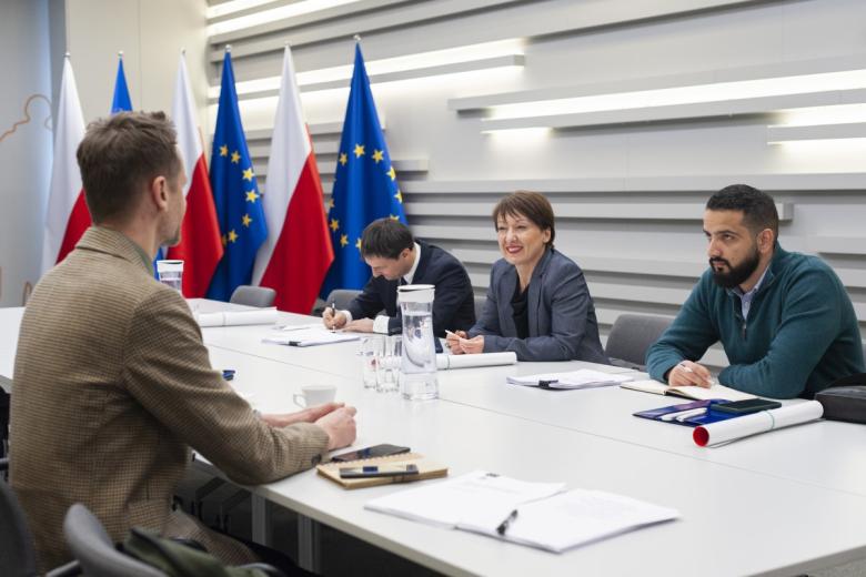 Pięć osób siedzi po dwóch stronach stołu i rozmawia, w tle flagi Polski i UE