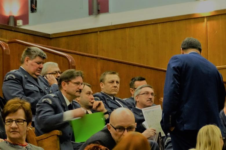 zdjęcie: na sali siedzi kilku mężczyzn w mundurach przed nimi stoi mężczyzna w garniturze