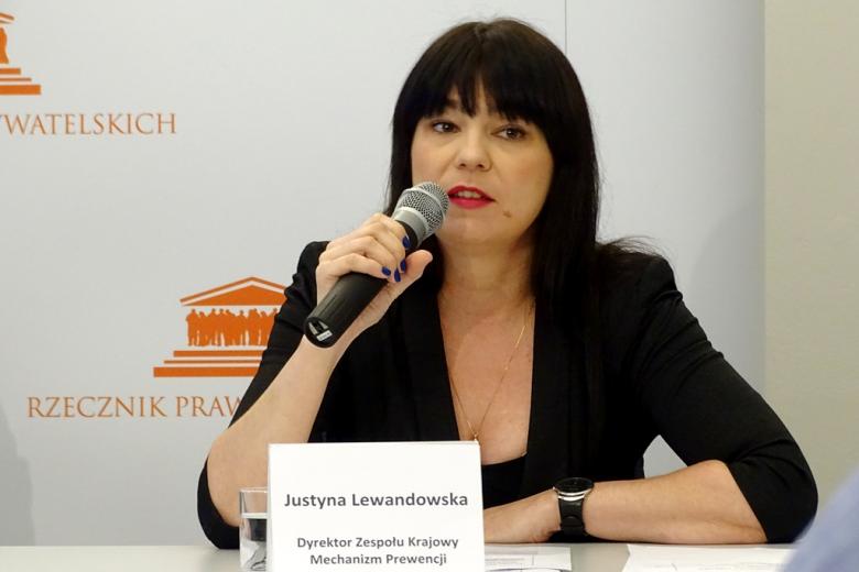 zdjęcie: kobieta w czarnej sukience i czrnym żakiecie siedzi za białym stołem konferencyjnym i mówi do mikrofonu