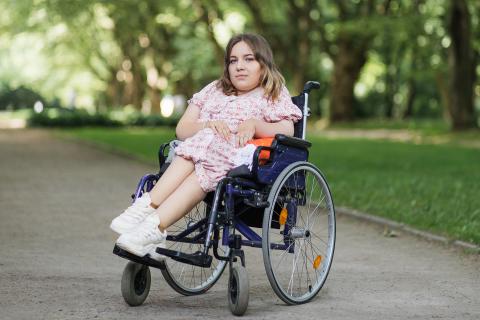Młoda kobieta z rdzeniowym zanikiem mięśni uśmiecha się siedząc na wózku w parku