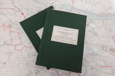 Dwie grube, zielone księgi z napisem "Plan urządzania lasu" leżące na mapie