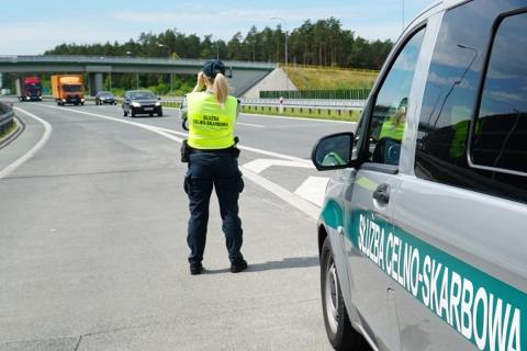 Funkcjonariuszka w mundurze Służby Celno-Skarbowej stoi przy służbowym samochodzie przy drodze szybkiego ruchu
